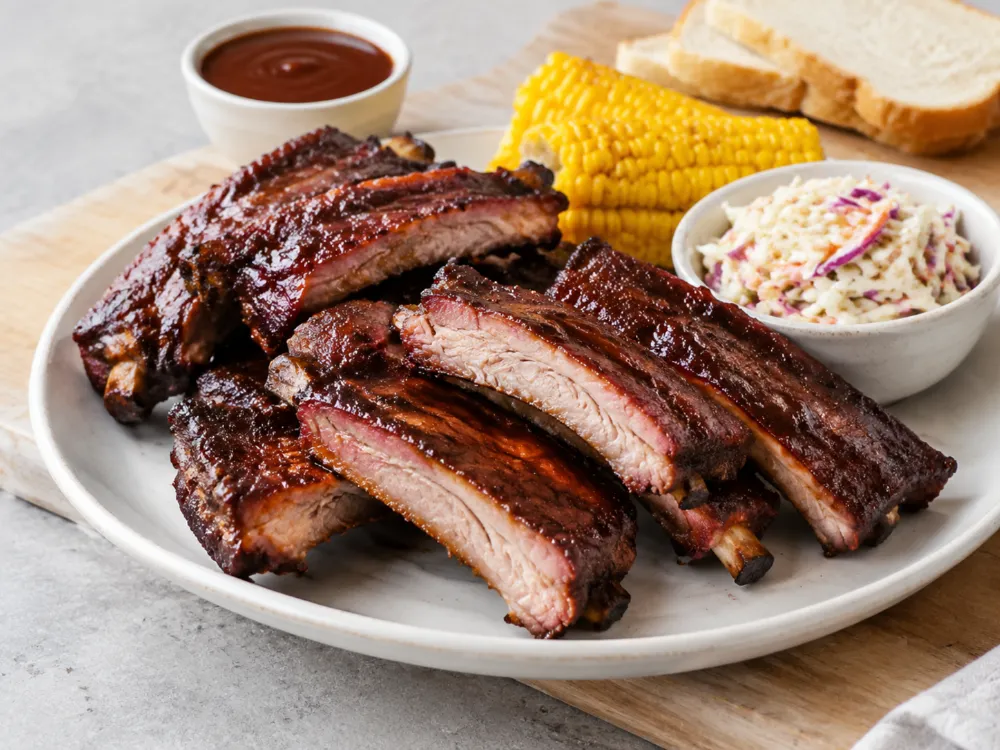 Żeberka BBQ pokrojone na pojedyncze kości na talerzu z surówką coleslaw, kukurydzą, pieczywem i sosem BBQ